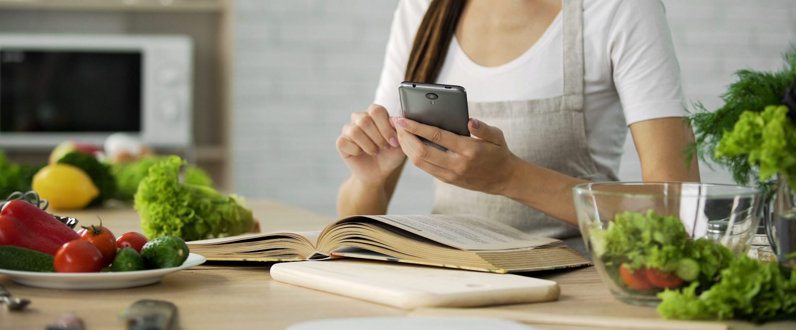 Woman learning on her phone and from a book in the kitchen / Photo Credit: Motortion Films / Shutterstock